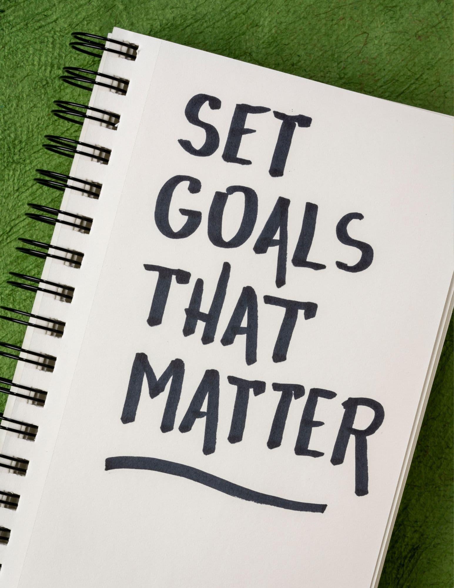 Spiral notebook on green background with handwritten text saying 'SET GOALS THAT MATTER' underlined.